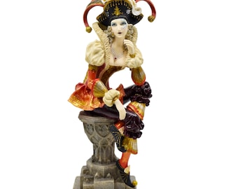 Op de afbeelding: Een beeldje van een vrouw gekleed als nar, met een rood, goud en zwart kostuum en een grote, veelkleurige hoed. Ze zit op een stenen voetstuk met haar hand op haar kin, denkend.