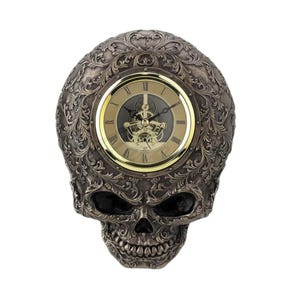 Könnte beinhalten: Eine bronzefarbene Schädel-Wanduhr mit einem verzierten Design und einem sichtbaren Uhrwerk. Das Zifferblatt der Uhr hat römische Ziffern und einen goldfarbenen Rand.