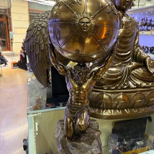 May include: A bronze statue of Atlas, a muscular figure, kneeling and supporting a globe. The globe features a sun design and zodiac symbols. A large Buddha statue is in the background. The statue is displayed on a glass shelf.
