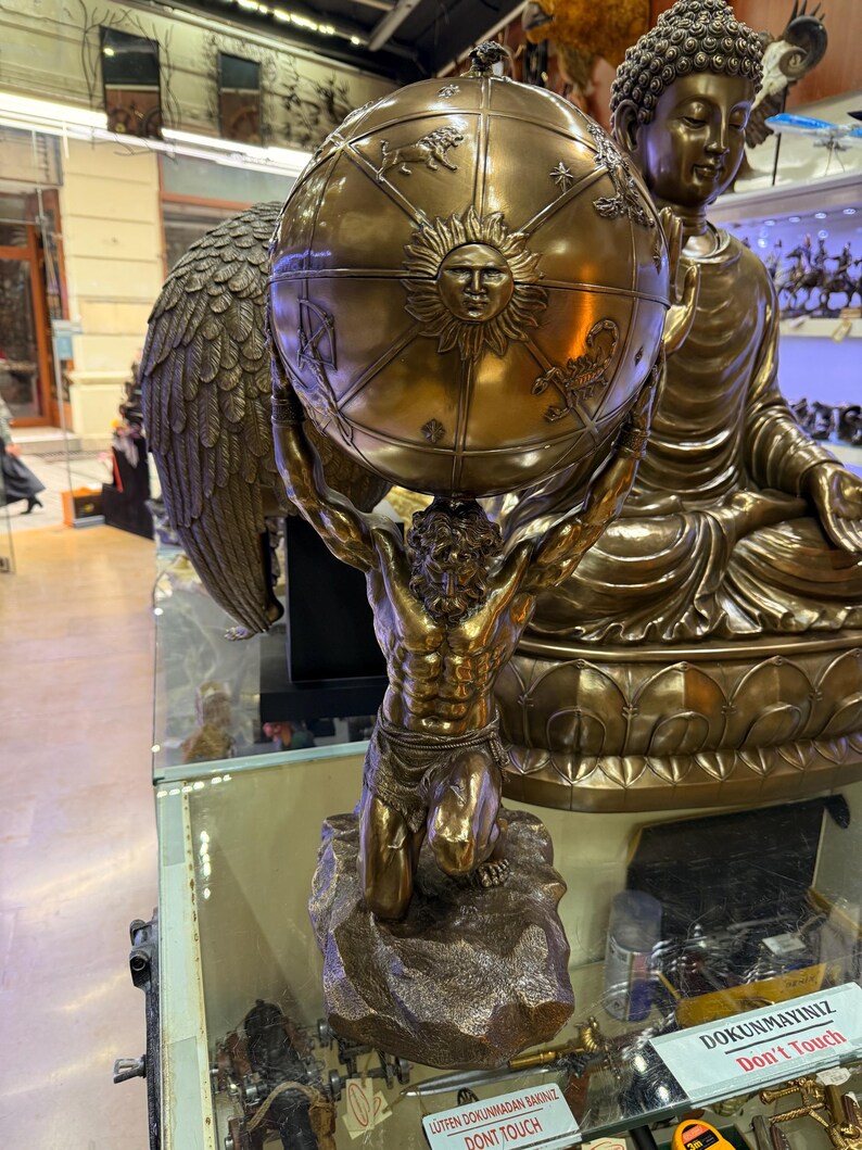 May include: A bronze statue of Atlas, a muscular figure, kneeling and supporting a globe. The globe features a sun design and zodiac symbols. A large Buddha statue is in the background. The statue is displayed on a glass shelf.