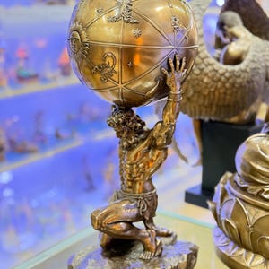 May include: A bronze Atlas statue kneeling and supporting a detailed globe. The globe features zodiac symbols and celestial designs. The statue is on a rocky base, showcasing intricate craftsmanship. The overall theme is classical art and mythology.