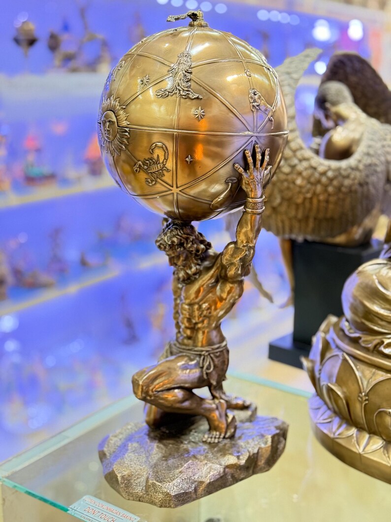 May include: A bronze Atlas statue kneeling and supporting a detailed globe. The globe features zodiac symbols and celestial designs. The statue is on a rocky base, showcasing intricate craftsmanship. The overall theme is classical art and mythology.