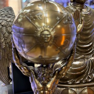 May include: A bronze sculpture of Atlas holding a globe, with detailed sun and zodiac designs. The globe is held aloft by a muscular figure. In the background, a bronze Buddha statue is visible. The overall theme is classical and spiritual.