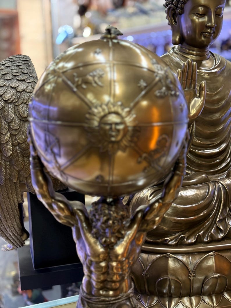May include: A bronze sculpture of Atlas holding a globe, with detailed sun and zodiac designs. The globe is held aloft by a muscular figure. In the background, a bronze Buddha statue is visible. The overall theme is classical and spiritual.
