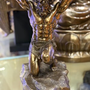 May include: A bronze sculpture of Atlas, a muscular male figure, kneeling and supporting a large sphere overhead. The sculpture has a detailed texture and is mounted on a rocky base. The figure's skin has a polished, reflective surface.