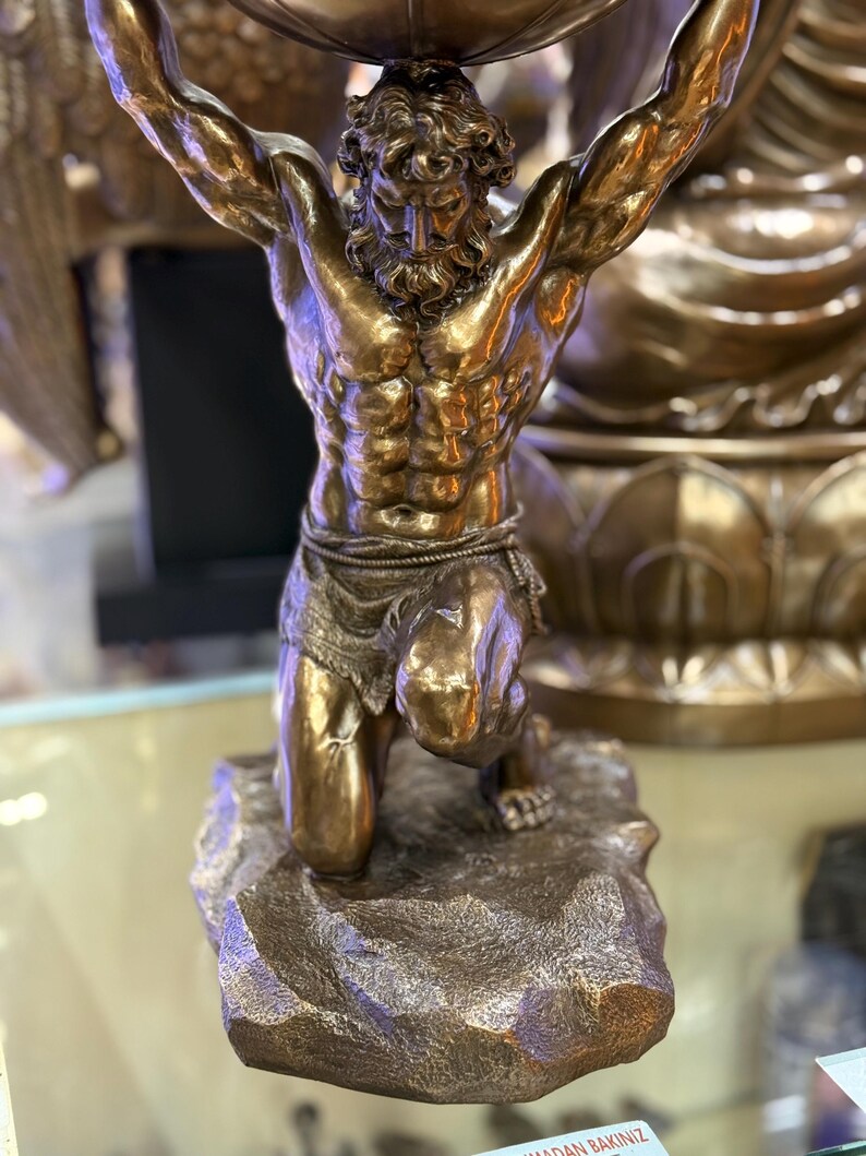 May include: A bronze sculpture of Atlas, a muscular male figure, kneeling and supporting a large sphere overhead. The sculpture has a detailed texture and is mounted on a rocky base. The figure's skin has a polished, reflective surface.