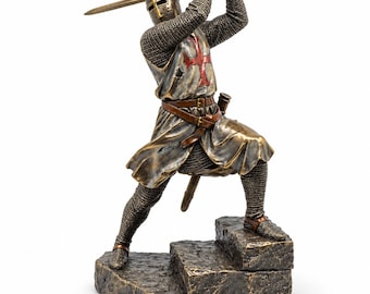 May include: A bronze-toned figurine of a medieval knight in chainmail armor, wearing a surcoat with a red cross emblem. The knight stands on stone steps, holding a sword raised above the helmeted head with both hands, ready for a strike.