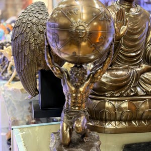 May include: A bronze statue of Atlas kneeling and supporting a globe. The globe features zodiac symbols and a sun face. A bronze Buddha statue is in the background. The statue is on display in a store.