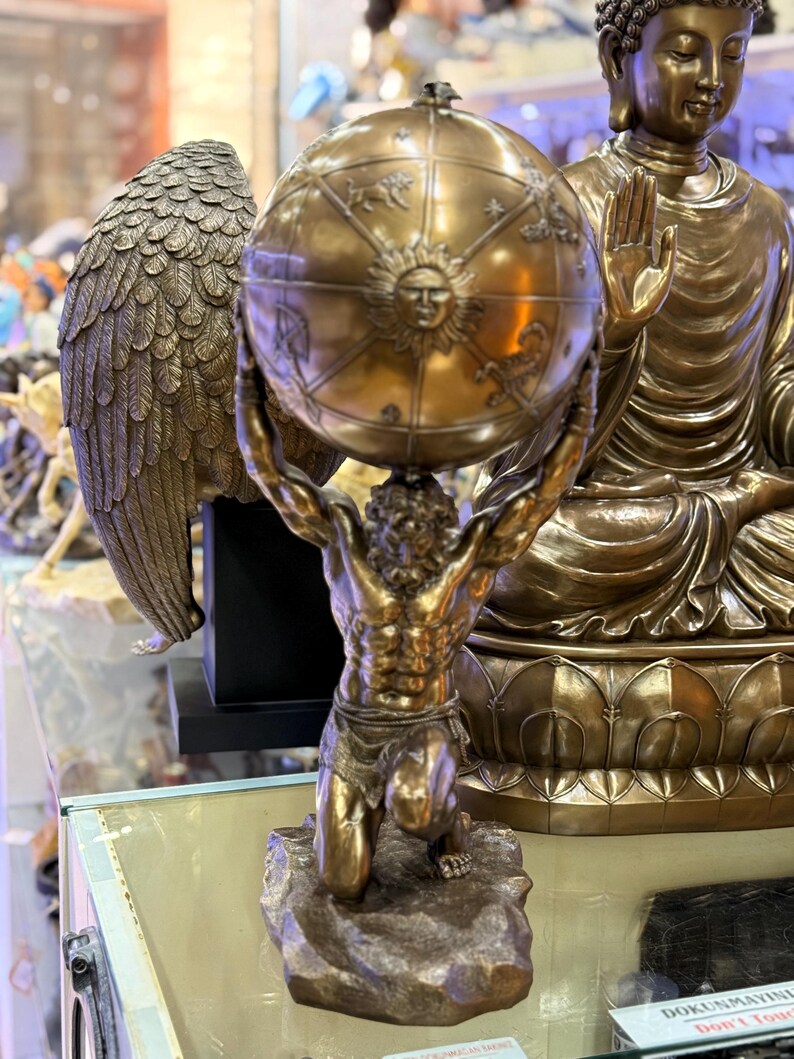 May include: A bronze statue of Atlas kneeling and supporting a globe. The globe features zodiac symbols and a sun face. A bronze Buddha statue is in the background. The statue is on display in a store.