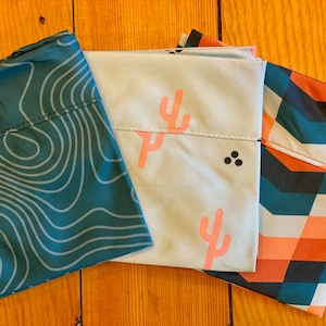 May include: Three fabric reusable shopping bags with different patterns. The first bag is teal with a white topographic map design. The second bag is light blue with a pink cactus design and three black dots. The third bag is a teal, orange, and white geometric pattern.