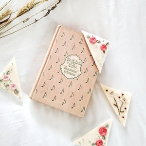 May include: Three white felt triangle bookmarks with embroidered floral designs. The bookmarks are on a white surface with a book titled "Invitation to the Waltz" by Rosamond Lehmann.