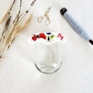 May include: A white fabric jar lid with a red and blue fruit print and white lace trim. The lid is on top of a clear glass jar.