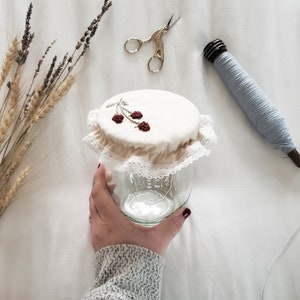 Op de afbeelding: Een wit stoffen deksel voor een pot met een kantversiering en geborduurde rode kersen. Het deksel zit op een glazen pot.