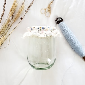 May include: A clear glass jar with a white lace and floral fabric lid. The jar is sitting on a white surface with a spool of blue thread and a pair of gold scissors.