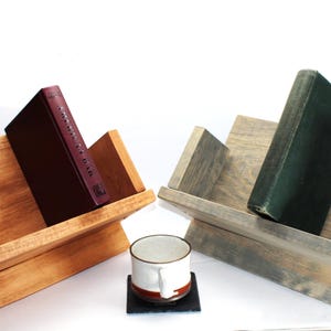 May include: Two wooden book stands, one stained light brown and the other gray, each holding a book. A maroon book sits in the brown stand, and a green book in the gray stand. A mug and coaster are in front.