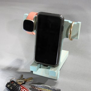 May include: A light blue wooden phone stand with a black phone, a pink and gold smartwatch, and a gold metal ring. There are also keys and a small toy fish on the table.
