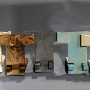 May include: Five wooden phone stands in different colors: natural, dark brown, gray, light blue, and natural. The stands are all the same design, with a T-shaped base and a rectangular top.
