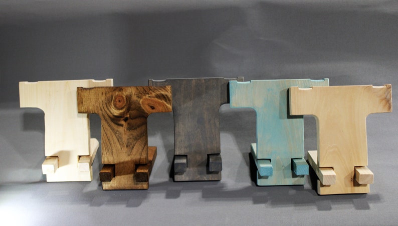 May include: Five wooden phone stands in different colors: natural, dark brown, gray, light blue, and natural. The stands are all the same design, with a T-shaped base and a rectangular top.