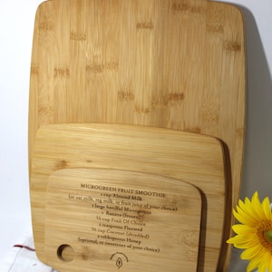 Puede incluir: Dos tablas de cortar de madera con una receta de batido de frutas y microgreens grabada en la tabla más pequeña. La receta incluye ingredientes como leche de almendras, microgreens, plátano, fruta de su elección, semillas de lino, coco y miel.
