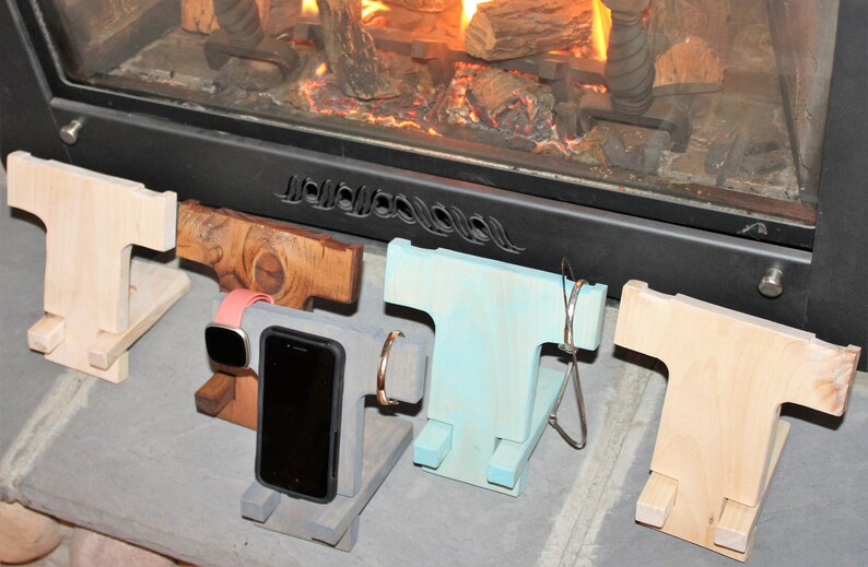 May include: Four wooden phone stands in different colors and styles. The stands are designed to hold a phone, watch, and other small items. The stands are arranged in a row on a gray surface.