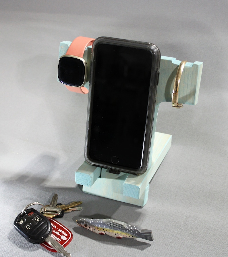 May include: A light blue wooden phone stand with a black phone, a pink and gold smartwatch, and a gold metal ring. There are also keys and a small toy fish on the table.