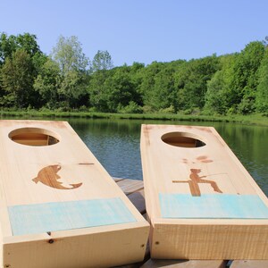 May include: Two wooden cornhole boards with a lake scene painted on them. One board has a silhouette of a fish jumping out of the water, and the other has a silhouette of a person fishing. The boards are sitting on a wooden deck in front of a lake.
