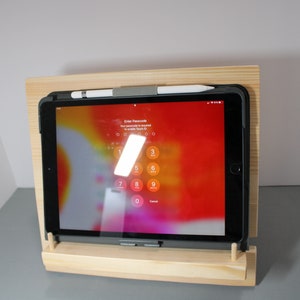 May include: A wooden stand with a black tablet in a protective case. The tablet is displaying a passcode screen with the text "Enter Passcode". A white stylus is resting on the top of the stand.