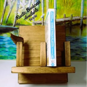 May include: A rustic wooden bookcase with a single shelf and two side supports. A blue book titled "Edible Wild Plants" is displayed on the shelf. The text "Rustic Bookcase Available Now" is written in white on a green and blue background.