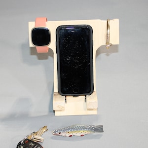 May include: A wooden phone stand with a slot for a phone, a watch holder, and a hook for keys. The stand is made of light wood and has a natural finish. A small toy fish is in the foreground.