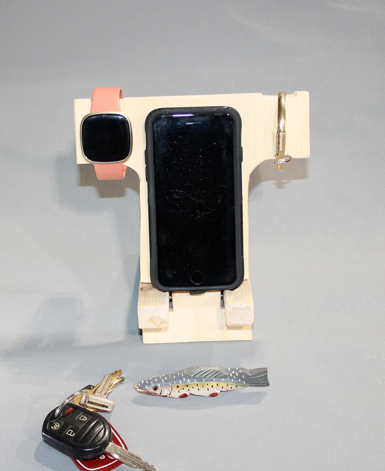May include: A wooden phone stand with a slot for a phone, a watch holder, and a hook for keys. The stand is made of light wood and has a natural finish. A small toy fish is in the foreground.