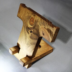 May include: A wooden phone stand with a natural wood finish. The stand is made of two pieces of wood that are joined together to form a T-shape. The stand is designed to hold a phone upright.
