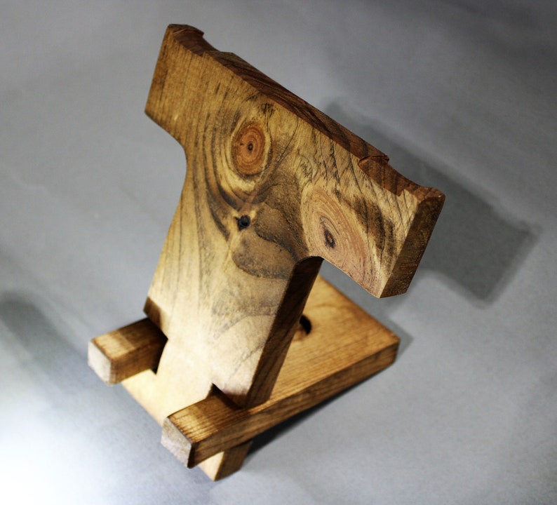May include: A wooden phone stand with a natural wood finish. The stand is made of two pieces of wood that are joined together to form a T-shape. The stand is designed to hold a phone upright.