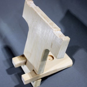 May include: A wooden phone stand with a simple design. The stand is made of light wood and has a cutout for a phone. The stand is folded up and ready to be used.