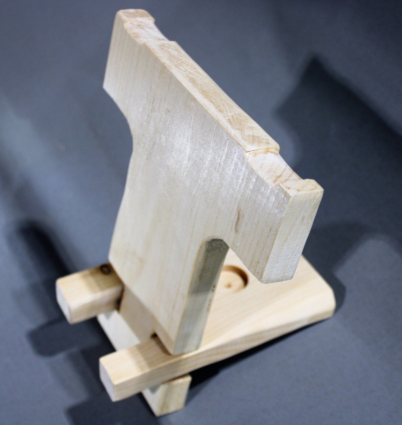 May include: A wooden phone stand with a simple design. The stand is made of light wood and has a cutout for a phone. The stand is folded up and ready to be used.
