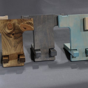 May include: Five wooden phone stands in different colors: natural, brown, gray, blue, and light wood. The stands are all the same design, with a T-shaped base and two legs that fold out to create a stand.