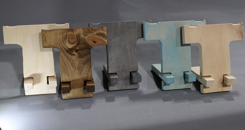 May include: Five wooden phone stands in different colors: natural, brown, gray, blue, and light wood. The stands are all the same design, with a T-shaped base and two legs that fold out to create a stand.