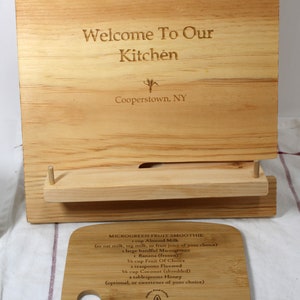 May include: A wooden cutting board with a recipe for a microgreen fruit smoothie engraved on it. The recipe includes ingredients like almond milk, microgreens, banana, fruit of choice, flaxseed, coconut, and honey. The board also has the text "Welcome To Our Kitchen Cooperstown, NY" engraved on it.