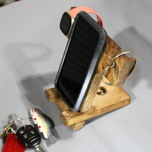 May include: A wooden phone stand with a slot for a phone, a watch, and a ring. The stand is made of natural wood with a rustic finish.