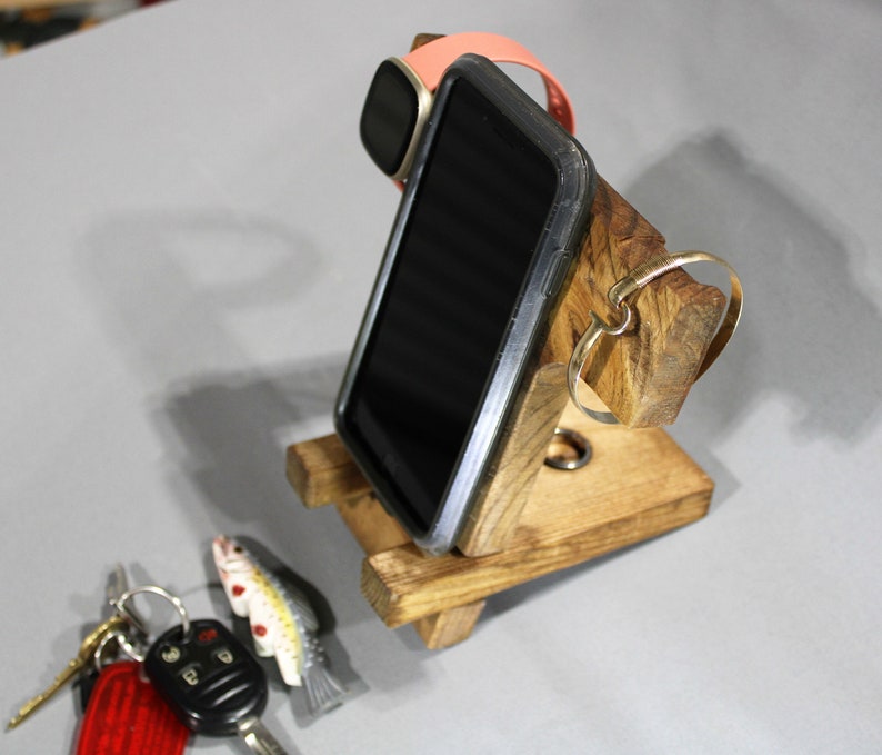 May include: A wooden phone stand with a slot for a phone, a watch, and a ring. The stand is made of natural wood with a rustic finish.
