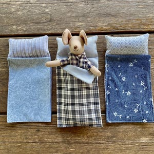 May include: Three blue and white patterned sleeping bags for dolls. One sleeping bag has a plaid pattern, another has a paisley pattern, and the third has a small floral pattern. A small stuffed mouse is lying in the plaid sleeping bag.