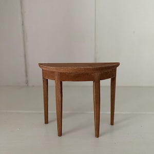 May include: A small, wooden, half-moon shaped table with three legs. The table is made of light brown wood and has a smooth finish.