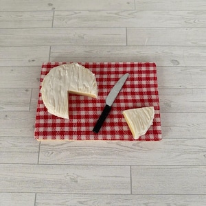 May include: A round of white cheese with a slice removed, presented on a red and white checked cutting board. A small silver knife with a black handle and a cheese wedge are also on the board. The board is on a light wood surface.