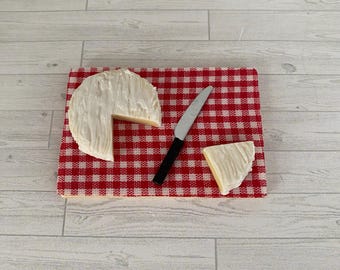 May include: A wheel of white cheese with a wedge cut out, placed on a red and white checkered cutting board. A small silver knife with a black handle and a cheese wedge are also on the board. The board is on a light wood surface.