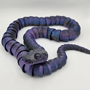 May include: A segmented snake sculpture in shades of purple and blue. The head is a darker gray with detailed scales and a single eye. The body is made of connected segments, creating a flexible, articulated form.