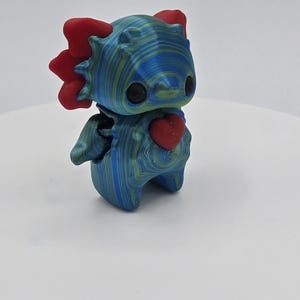 May include: A 3D-printed figurine of a stylized dragon with a heart on its chest. The dragon is primarily blue with green accents, and red accents on its head and heart. The figurine has large black eyes and small wings.