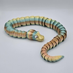 Może przedstawiać: Zabawka w kształcie węża, elastyczna i segmentowana, w kolorze zielonym, złotym i brązowym. Wąż jest zwinięty w k&oacute;łko, głową zwr&oacute;coną w stronę widza. Głowa ma zębaty uśmiech i duże, czarne oczy.