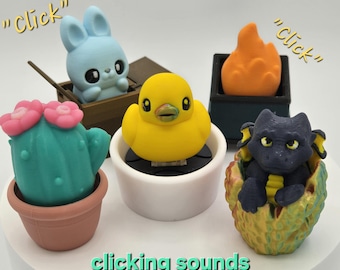 May include: A collection of colorful, whimsical figurines, including a blue bunny in a wooden cart, a yellow duck, a cactus with pink flowers, an orange flame, and a dark blue dragon in an egg. The text "clicking sounds" is at the bottom.