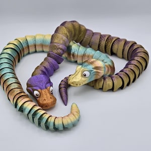 May include: Two colorful, segmented, snake-like toys with cartoon faces. The snake on the left is purple and green, while the snake on the right is gold and green.