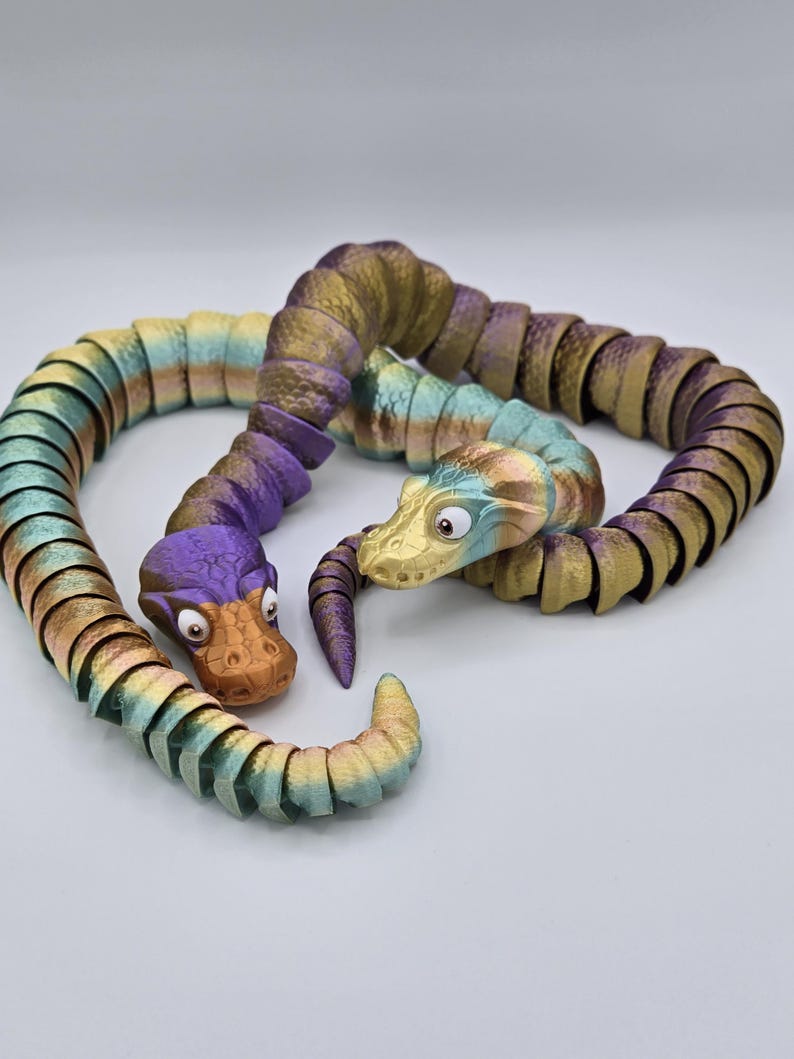 Może przedstawiać: Dwie kolorowe, segmentowane zabawki w kształcie węża z kresk&oacute;wkowymi twarzami. Wąż po lewej stronie jest fioletowy i zielony, a wąż po prawej stronie jest złoty i zielony.