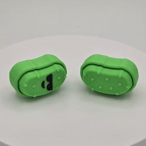 May include: Two green pickle-shaped erasers. One eraser has a black letter "B" on its side. The erasers have a bumpy texture and are set against a white background. The erasers are a fun and unique stationery item.